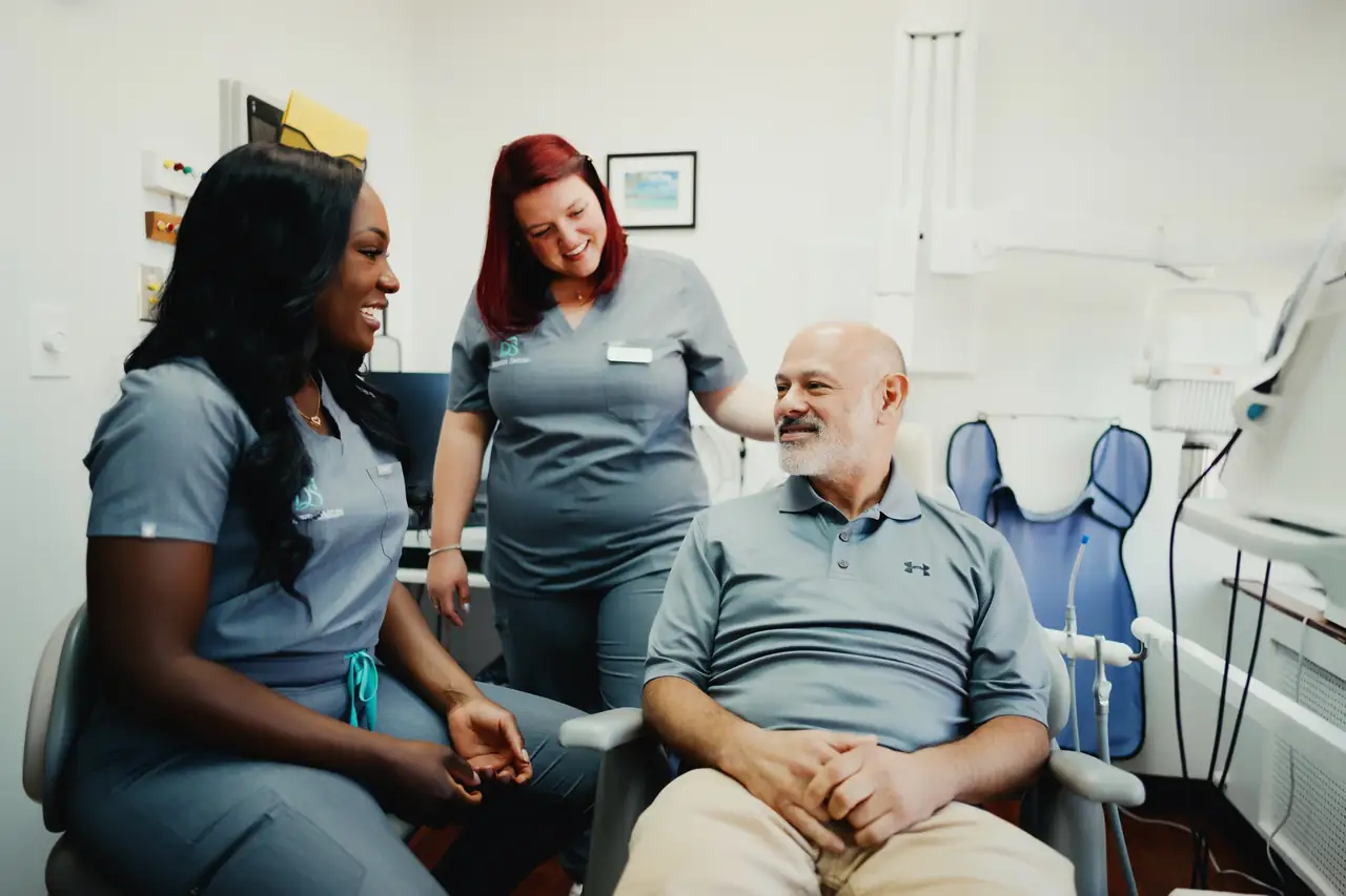 Two Designer Smiles team members in grey scrubs talking with a smiling patient in a treatment room.