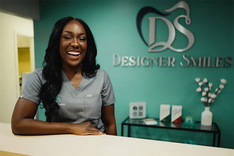 A smiling team member at the Designer Smiles front desk, the teal logo wall behind her.