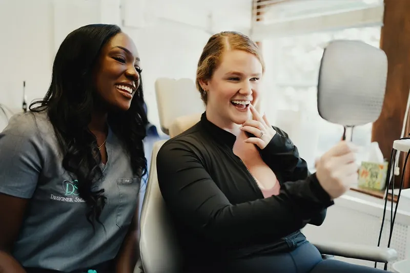 A smiling patient checking her smile in a hand mirror beside a team member in scrubs.
