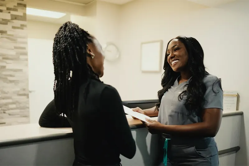 A team member in grey scrubs greeting a patient at the front desk.